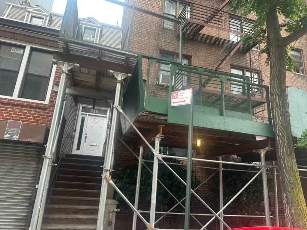 Storefront building with metal stairway and green suspended sidewalk scaffolding in front of a brick apartment, showcasing Suspended Scaffolding Services In Bronx for exterior construction and maintenance work.